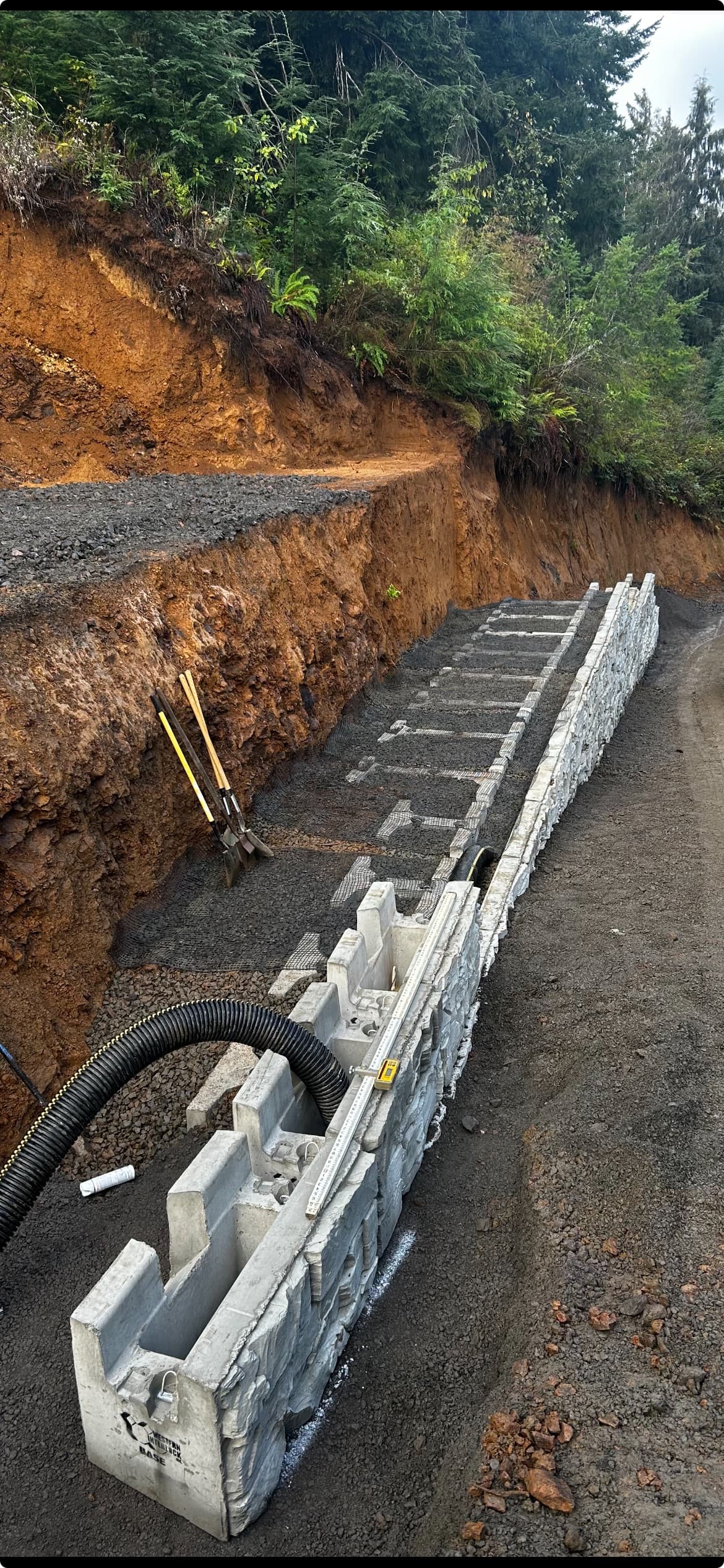 Retaining wall base course set in the excavated trench with drainage pipe against the steep cut bank at a hillside property in Otis, Oregon